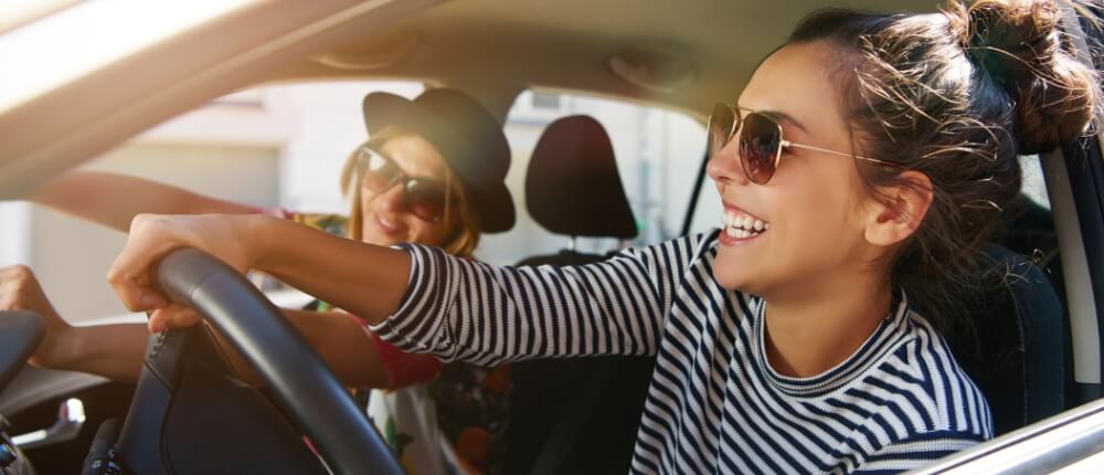 Zwei junge Frauen in einer Sonnenbrille fahren in einem Auto und freuen sich über ihre günstige Autoversicherung.