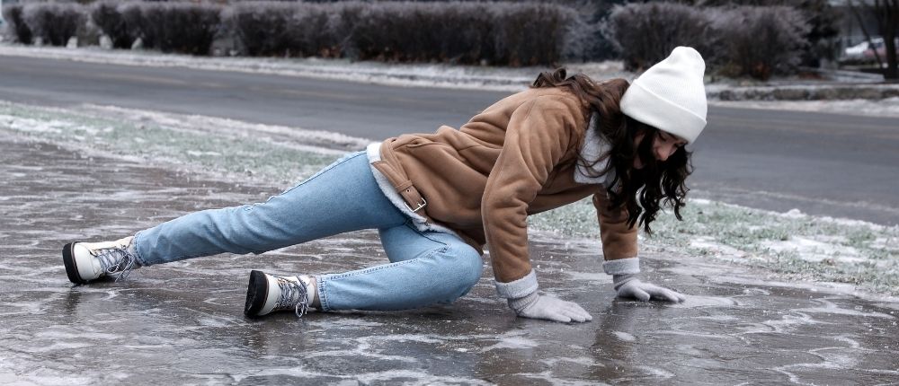 Eine Frau mit Hut und Handschuhen ist auf einer verschneiten, glatten Straße zu Boden gestürzt.